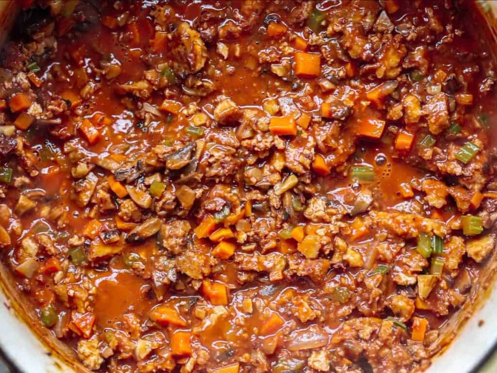 A close-up of hearty vegan bolognese simmering in a pot, with visible pieces of plant-based meat, diced carrots, celery, and a rich tomato base.